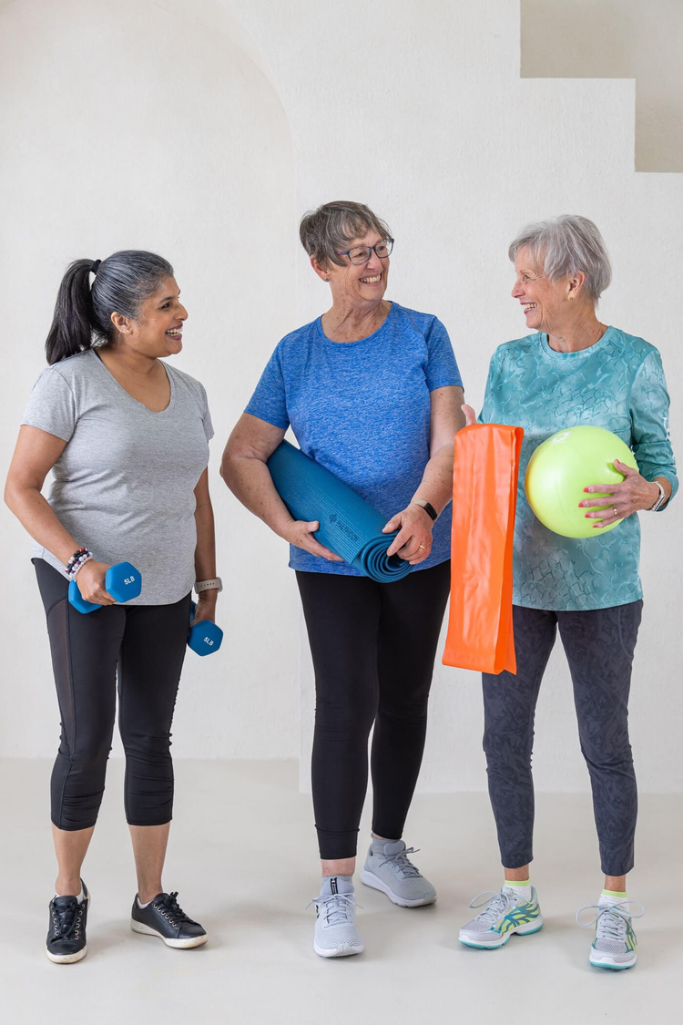 Three older women chatting with exercise bands, pilates ball and weights.